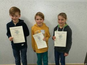 Drei Kinder stehen nebeneinander und halten jeweils ein Zertifikat in der Hand. Sie lächeln in die Kamera. Im Hintergrund ist eine helle Wand zu sehen.