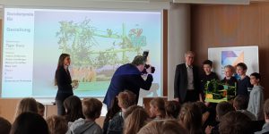 Eine Gruppe von Schülern steht auf der Bühne und präsentiert ein Projekt. Im Hintergrund ist eine Präsentationsfolie mit einer grünen Landschaft zu sehen. Ein Mann fotografiert die Schüler, während ein anderer Mann und eine Frau aufmerksam zuhören. Im Vordergrund sitzen mehrere Kinder im Publikum.