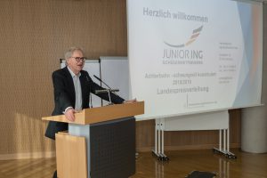 Ein Mann steht an einem Rednerpult und spricht. Im Hintergrund ist eine Präsentation mit dem Text 
