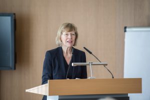 Eine Frau steht am Rednerpult und hält eine Ansprache. Sie trägt einen schwarzen Blazer und hat blonde Haare. Im Hintergrund sind ein Fernseher und ein Flipchart zu sehen. Die Umgebung wirkt modern und hell.