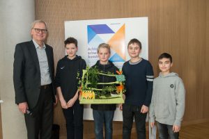Eine Gruppe von vier Schülern steht zusammen und präsentiert ein kreatives Projekt. Sie halten ein Modell, das mit Pflanzen und bunten Elementen gestaltet ist. Neben ihnen steht ein älterer Mann in formeller Kleidung. Im Hintergrund ist ein Plakat mit geometrischen Formen zu sehen.