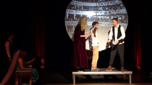 Eine Theateraufführung mit mehreren Darstellern auf einer Bühne. Im Vordergrund stehen zwei Personen im Gespräch, während eine dritte Person im Hintergrund sitzt. Eine große runde Projektionsfläche zeigt eine historische Theateransicht. Die Atmosphäre ist dramatisch, mit einem roten und schwarzen Bühnenhintergrund.