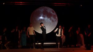 Eine Theateraufführung mit mehreren Schauspielern auf der Bühne. Im Hintergrund ist eine große, leuchtende Mondprojektion zu sehen. Ein Schauspieler in einem schwarzen Anzug steht im Vordergrund und posiert, während die anderen Darsteller in verschiedenen Kostümen hinter ihm stehen. Die Beleuchtung ist dramatisch und erzeugt eine stimmungsvolle Atmosphäre.