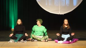 Drei Personen sitzen auf Matten in Meditationshaltung auf einer Bühne. Die linke Person hat dunkle Haare und trägt ein schwarzes Oberteil, die mittlere Person hat grüne Haut und einen muskulösen Körper, während die rechte Person ebenfalls dunkle Haare hat und ein helles Oberteil trägt. Im Hintergrund sind grüne Lichter und eine große runde Projektion zu sehen.
