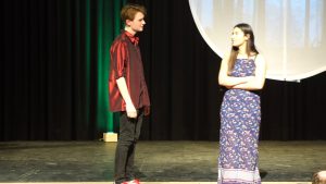 Zwei Schauspieler auf einer Bühne während einer Theateraufführung. Der männliche Schauspieler trägt ein rotes Hemd und schwarze Hosen, während die weibliche Schauspielerin ein langes, blaues Kleid mit Mustern trägt. Beide stehen sich gegenüber und scheinen in ein Gespräch vertieft zu sein. Im Hintergrund ist eine große, runde Leinwand und der Bühnenboden ist dunkel.
