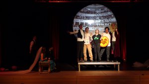 Eine Theateraufführung auf einer Bühne. Fünf Schauspieler stehen im Vordergrund, während ein aufwändiger Bühnenhintergrund mit einer großen Kulisse zu sehen ist. Eine Person sitzt links auf einer Bank und beobachtet die Szene. Die Schauspieler zeigen verschiedene Posen, einige halten Requisiten in den Händen. Die Beleuchtung ist stimmungsvoll und hebt die Darsteller hervor.