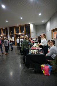 Eine Veranstaltung in einem modernen Raum mit großen Fenstern. An einem Tisch stehen mehrere Personen, die Produkte oder Informationen anbieten. Im Hintergrund sind weitere Teilnehmer der Veranstaltung zu sehen, die miteinander sprechen und sich bewegen. Der Boden ist dunkel und die Wände sind aus Beton, während die Decke hell erleuchtet ist.