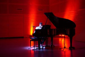 Ein junger Mann sitzt an einem Klavier, umgeben von rotem Licht. Er spielt und konzentriert sich auf das Instrument. Der Raum ist schwach beleuchtet, mit einer hellen Lichtquelle, die auf den Pianisten und das Klavier gerichtet ist.