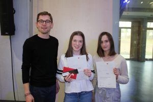 Drei Personen stehen in einem hellen Raum. Ein Mann auf der linken Seite trägt ein schwarzes Oberteil und lächelt. Neben ihm stehen zwei junge Frauen, die jeweils ein Dokument und Geschenkkarten in der Hand halten. Die Frauen tragen helle Oberteile und sehen ebenfalls erfreut aus. Im Hintergrund sind große Fenster und eine Bühne zu erkennen.