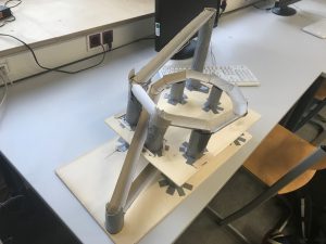 Modell einer Konstruktion aus Papier und Karton auf einem Tisch. Die Struktur zeigt eine Plattform mit mehreren Säulen und Bögen, die eine kreisförmige Form bilden. Im Hintergrund sind ein Computer und eine Tastatur sichtbar.