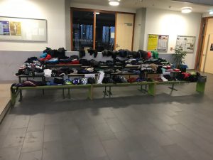 Ein Tisch mit verschiedenen Kleidungsstücken ist in einem Raum aufgestellt. Die Kleidung ist ordentlich aufgereiht, darunter Pullover, T-Shirts und Hosen. Der Raum hat große Fenster mit Tageslicht und eine klare, moderne Gestaltung.