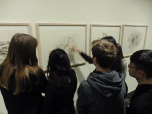 Eine Gruppe von fünf Personen steht vor einer Wand mit gerahmten Zeichnungen. Eine Person zeigt mit dem Finger auf eine der Zeichnungen, während die anderen aufmerksam zuschauen. Alle Personen tragen dunkle Kleidung und scheinen an der Kunst interessiert zu sein.