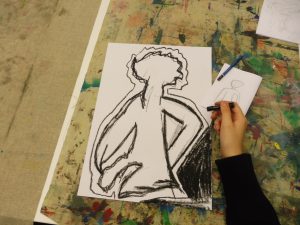 Eine Person zeichnet mit einem schwarzen Stift auf einem großen weißen Blatt Papier, das eine stilisierte Figur mit lockigem Haar zeigt. Im Hintergrund sind bunte Farbspritzer auf einer Tischoberfläche sichtbar. Neben dem großen Blatt liegt ein kleineres Blatt mit einer einfachen Skizze.
