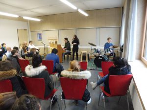 Eine Gruppe von Personen sitzt auf roten Stühlen in einem Raum. Auf der Bühne spielen zwei Musiker: eine Person am Klavier und eine Sängerin. Neben ihnen steht ein Schlagzeuger. Im Hintergrund sind weitere Zuhörer sichtbar, die aufmerksam der Darbietung folgen. Der Raum hat große Fenster, durch die Tageslicht hereinfällt.