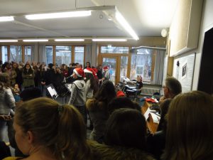 Eine große Gruppe von Menschen steht in einem Raum mit Fenstern, die auf einen Außenbereich blicken. Einige Personen tragen Weihnachtsmützen. Im Vordergrund sind die Rücken von mehreren Personen zu sehen, während eine kleine Gruppe von Menschen mit Notenblättern auf einer Bühne oder einem erhöhten Bereich steht. Der Raum wirkt festlich und ist gut beleuchtet.