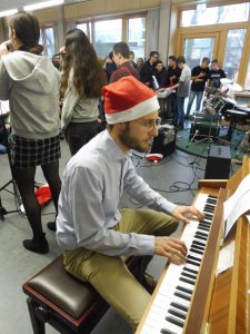 Ein Mann mit einer roten Weihnachtsmütze sitzt an einem Klavier und spielt. Im Hintergrund sind mehrere Personen zu sehen, die an einer musikalischen Aufführung teilnehmen. Die Umgebung ist hell erleuchtet und wirkt festlich.