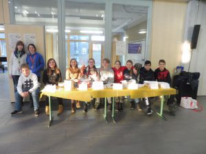 Eine Gruppe von Jugendlichen sitzt an einem Tisch mit gelber Tischdecke. Auf dem Tisch stehen verschiedene Behälter und Karten. Im Hintergrund sind Fenster und eine Wand zu sehen. Einige Jugendliche stehen hinter dem Tisch, während andere sitzen. Die Szene vermittelt eine freundliche, gemeinschaftliche Atmosphäre.