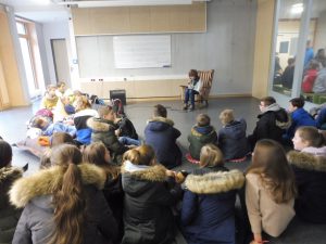 Eine Gruppe von Kindern sitzt auf dem Boden in einem Klassenraum und hört einer Person zu, die auf einem Stuhl sitzt und liest. Die Kinder tragen warme Jacken mit Kapuzen und haben lange Haare. Im Hintergrund ist eine Tafel zu sehen, und der Raum hat große Fenster.