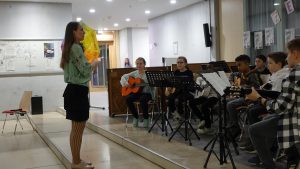 Eine Musiklehrerin steht vorne und dirigiert eine Gruppe von Schülern, die mit Gitarre und anderen Instrumenten spielen. Der Raum hat große Fenster und eine helle Beleuchtung, im Hintergrund sind Notenständer und ein paar Zeichnungen an der Wand.