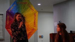 Eine Frau spricht mit einem Mikrofon, während sie vor einem bunten Regenschirm steht, der verschiedene Worte in unterschiedlichen Farben zeigt. Im Vordergrund sitzt ein Mädchen und hört aufmerksam zu. Der Hintergrund ist hell und neutral.