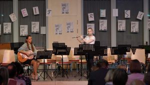Zwei Musikerinnen auf einer Bühne während einer Aufführung. Eine spielt Gitarre, die andere Flöte. Im Hintergrund sind Notenständer und Plakate mit dem Schriftzug „ZEITREISE“ zu sehen. Das Publikum sitzt vor der Bühne und hört zu.
