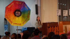 Eine Frau sitzt an einem Klavier und spielt. Sie hat lange Haare und trägt ein helles Oberteil. Hinter ihr steht ein großer Regenbogen-Schirm mit verschiedenen Farben und Schriftzügen. Der Raum ist mit Menschen gefüllt, die ihr aufmerksam zuhören. Im Hintergrund sind einige Plakate an der Wand sichtbar.