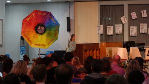 Eine junge Frau steht an einem Podium in einem Raum, umgeben von einem Publikum. Im Hintergrund steht ein großer, farbenfroher Regenschirm in Regenbogenfarben. An der Wand hängen mehrere Papierarbeiten oder Plakate. Das Licht ist gedämpft, und die Zuschauer sind aufmerksam auf die Präsentation gerichtet.