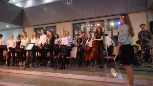 Eine Gruppe von Musikern steht auf einer Bühne in einem Innenraum. Die Musiker tragen überwiegend weiße Hemden und schwarze Hosen oder Röcke. Im Vordergrund ist eine Dirigentin zu sehen, die mit dem Rücken zur Kamera steht. Im Hintergrund sind Instrumente wie Geigen und ein Schlagzeug zu erkennen. Die Wände sind mit bunten Plakaten dekoriert, und es gibt eine helle Beleuchtung.