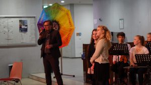 Eine Person spricht am Mikrofon auf einer Bühne, während eine andere Person in der Nähe steht. Im Hintergrund sind mehrere Musikerinnen und Musiker mit Notenständern zu sehen. Ein bunter Regenschirm mit Regenbogenfarben ist ebenfalls im Bild, was eine festliche Atmosphäre vermittelt.