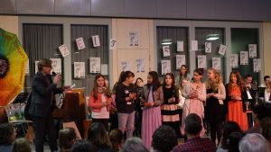 Eine Gruppe von Kindern und Jugendlichen steht auf einer Bühne und präsentiert etwas. Ein Erwachsener spricht in ein Mikrofon. Im Hintergrund sind Instrumente und eine bunte Dekoration zu sehen. An den Wänden hängen Zeichnungen oder Plakate. Das Publikum sitzt im Vordergrund und schaut aufmerksam zu.
