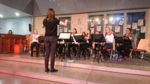 Eine Musikgruppe sitzt auf einer Bühne und spielt Instrumente. Eine Person steht vorne und dirigiert. Im Hintergrund ist ein Klavier zu sehen, und die Wände sind mit Zeichnungen dekoriert. Die Musikerinnen sind in formeller Kleidung und haben Notenständer vor sich.