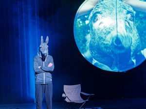 Ein Darsteller steht auf einer Bühne und trägt eine tierähnliche Maske. Im Hintergrund ist ein großer Bildschirm, der eine Nahaufnahme eines Tieres zeigt. Die Szenerie ist in bläuliches Licht getaucht, und es gibt einen leeren Stuhl auf der Bühne.