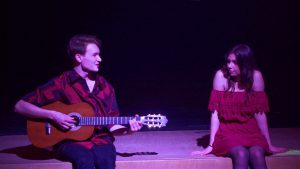 Ein junger Mann spielt Gitarre und singt auf einer Bühne, während eine junge Frau in einem roten Kleid auf einem Podest sitzt und ihn ansieht. Die Szene ist schwach beleuchtet, was eine intime Atmosphäre schafft.