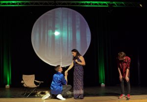 Eine Theateraufführung auf einer Bühne mit einem großen, runden Hintergrund, der einen Wald darstellt. Zwei Schauspieler stehen im Vordergrund: ein Mann in blauer Kleidung kniet vor einer Frau in einem langen Kleid und scheint sie um ihre Hand zu bitten. Ein dritter Schauspieler, der in einem karierten Hemd gekleidet ist, steht daneben und schaut zu. Im Hintergrund sind grüne Lichter zu sehen, die die Atmosphäre der Szene verstärken. Ein Stuhl aus Canvas ist ebenfalls im Bild.