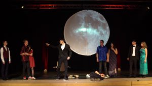 Eine Theateraufführung auf einer Bühne mit mehreren Darstellern in verschiedenen Kostümen. Im Hintergrund ist ein großer Mond abgebildet, der die Szene beleuchtet. Einige Darsteller stehen in verschiedenen Posen, während einer im Vordergrund gestikuliert. Ein Darsteller liegt auf der Bühne. Die Atmosphäre wirkt dramatisch und künstlerisch.