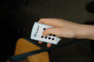 Eine Hand hält eine Fernbedienung mit mehreren Tasten. Der Hintergrund ist unscharf und zeigt einen Stuhl.
