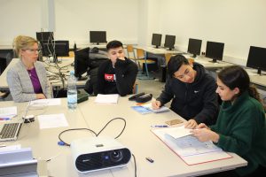 Eine Gruppe von vier Personen sitzt an einem Tisch in einem modernen Klassenraum. Auf dem Tisch befinden sich Laptops, Notizmaterialien und ein Projektor. Eine Lehrerin blickt auf die Schüler, die an einem Projekt arbeiten und in ihre Unterlagen vertieft sind. Im Hintergrund sind Computerarbeitsplätze zu sehen.