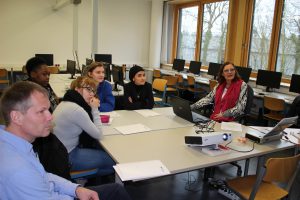 Eine Gruppe von sechs Personen sitzt an einem Tisch in einem Raum mit Computerarbeitsplätzen. Auf dem Tisch befinden sich Dokumente und ein Laptop. Eine Person spricht, während die anderen aufmerksam zuhören. Der Raum hat große Fenster, durch die Tageslicht fällt.