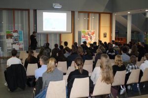 Eine Gruppe von Menschen sitzt in einem Seminarraum und schaut auf eine Präsentation, die an einer Leinwand gezeigt wird. Im Vordergrund steht eine Person, die spricht. Die Teilnehmer sind überwiegend junge Erwachsene, die in Reihen in Stühlen sitzen. Der Raum ist hell und modern gestaltet, mit verschiedenen Informationsständen im Hintergrund.