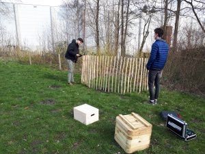 Zwei Personen arbeiten im Freien an einem Holzzaun. Die Umgebung ist grün mit Gras und einigen Bäumen im Hintergrund. Auf dem Boden liegen zwei Holzkisten und ein kleiner schwarzer Koffer.