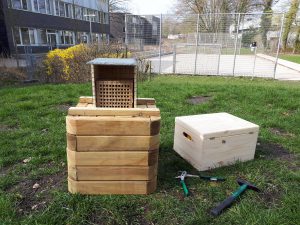 Holzkonstruktion bestehend aus einem quadratischen, höheren Behälter mit einem Gitterdeckel und einer niedrigeren, kastenförmigen Holzbox, die auf einer grünen Wiese steht. Im Vordergrund sind Werkzeuge wie ein Hammer und eine Zange zu sehen. Im Hintergrund sind einige Bäume und ein Zaun sichtbar.