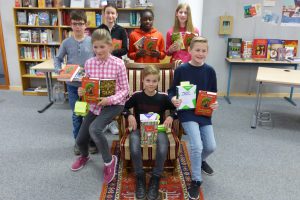 Sechs Kinder stehen und sitzen in einer Bibliothek, umgeben von Bücherregalen. Einige halten Bücher in den Händen, während ein Mädchen in einem Stuhl sitzt. Die Kinder lächeln und sind in Freizeitkleidung. Im Hintergrund sind Tische und weitere Bücher sichtbar.