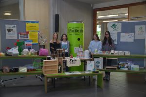 Drei Personen stehen vor einem Tisch mit verschiedenen Produkten und Informationen, umgeben von Plakaten und einem großen grünen Behälter. Der Tisch ist mit Holzregalen und verschiedenen Artikeln dekoriert, darunter Lebensmittel, Behälter und Info-Materialien. Im Hintergrund sind Wände mit weiteren Informationen und Plakaten zu sehen.