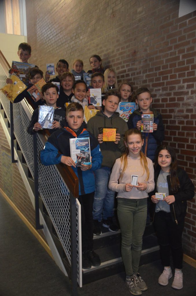 Eine Gruppe von 20 Kindern steht auf einer Treppe und hält verschiedene Gegenstände in den Händen. Die Kinder sind in zwei Reihen angeordnet, einige stehen im Hintergrund und andere im Vordergrund. Sie lächeln und schauen in die Kamera. Die Wand hinter ihnen ist aus Ziegelstein, und der Boden ist mit einem dunklen Material bedeckt. Das Licht im Raum ist hell.