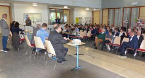 Eine Gruppe von Menschen sitzt in einem großen Raum, während vorne mehrere Personen an einem Tisch sprechen. Die Zuhörer sind auf Stühlen angeordnet und schauen aufmerksam in Richtung der Redner. Der Raum ist hell erleuchtet und hat große Fenster. Im Hintergrund sind Informationsplakate und eine Präsentation sichtbar.