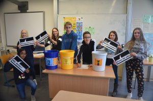 Sechs Schüler stehen an einem Tisch in einem Klassenzimmer und halten Schilder in den Händen. Auf dem Tisch stehen zwei Eimer, ein blauer und ein gelber, die mit verschiedenen Symbolen versehen sind. Im Hintergrund sind ein Whiteboard und Plakate an der Wand zu sehen.