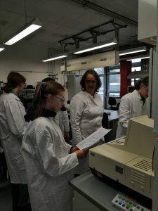 Eine Gruppe von Personen in weißen Laborkitteln steht in einem Labor. Eine Person mit einer Schutzbrille liest ein Dokument, während eine andere Person ihr zuhört. Im Hintergrund sind Laborausrüstungen und Geräte sichtbar. Die Atmosphäre wirkt konzentriert und arbeitsam.