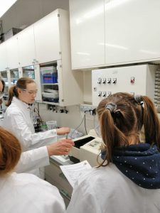 Zwei Personen in Laborkitteln stehen in einem Labor. Eine Person hält ein Glas und schaut auf ein Gerät, während die andere Person mit einem Blatt Papier in der Hand neben ihr steht. Im Hintergrund sind Laboreinrichtungen und Schränke sichtbar.