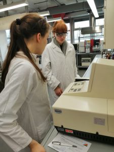 Zwei Personen in Laborkitteln arbeiten an einem Laborgerät. Eine Person schaut auf das Gerät, während die andere etwas notiert. Im Hintergrund sind Laborausrüstungen und Fenster sichtbar, die Tageslicht hereinlassen.