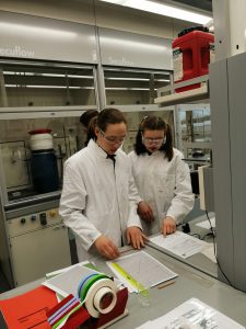 Zwei Personen in weißen Laborkitteln und Schutzbrillen stehen an einem Tisch in einem Labor. Sie schauen auf Dokumente und arbeiten zusammen. Im Vordergrund sind verschiedene farbige Bänder und Papierunterlagen zu sehen. Im Hintergrund sind Laborausrüstungen und Chemikalien in Behältern sichtbar.